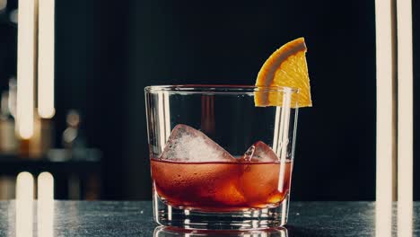 negroni cocktail garnished with orange slice sits on a bar counter, ice cubes melting slowly in the amber liquid, with blurred bar shelves in the background