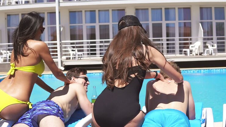 Trois hommes recevant un massage de jeunes femmes en bikini, allongés sur des transats au bord de la piscine dans une belle villa et buvant de la bière