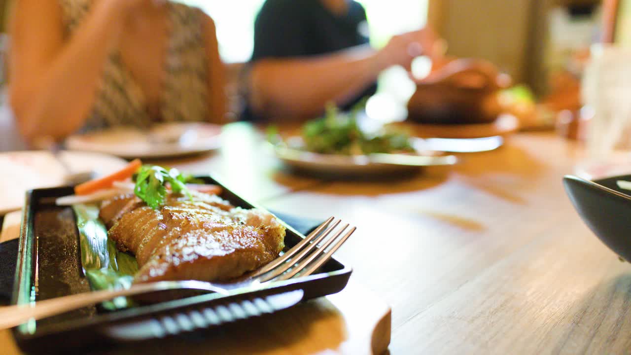Close-up of grilled pork neck dish, diners in background, natural daylight, shallow depth of field