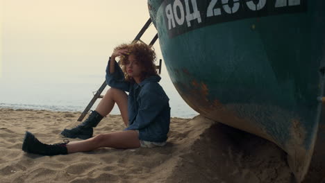 Sad-woman-sit-beach-near-old-boat-cloudy-morning.-Attractive-stylish-girl-posing