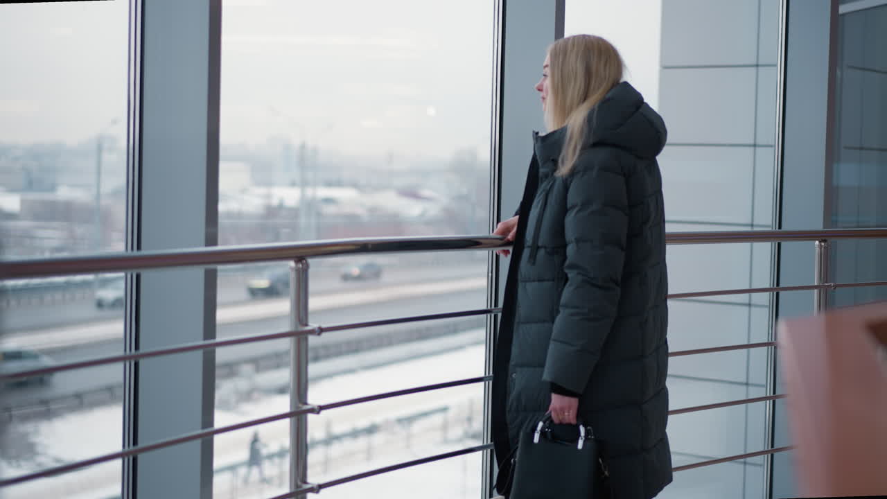 mujer con chaqueta negra de pie cerca de la ventana de vidrio, mirando hacia afuera mientras observa los coches que pasan, con el entorno urbano visible en el fondo