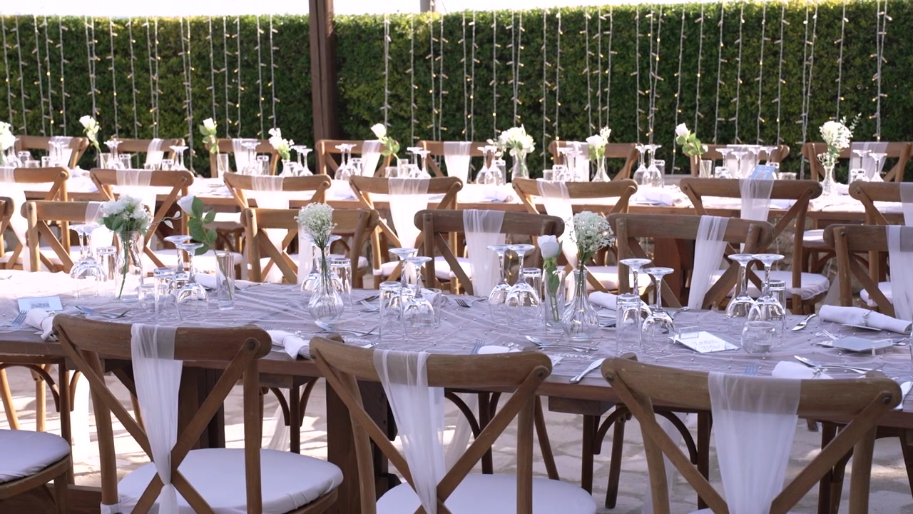 Borde er dækket op til et bryllup i en have. Hvert bord har blomster og glas. Lyskæder hænger over for at give lys. Opsætningen viser forberedelse til begivenheden.