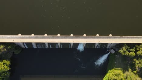 Luftaufnahme-Von-Oben-Nach-Unten-Vom-Dammbau-Mit-Wasserfall,-Der-Im-Sonnenlicht-In-Den-Fluss-Und-Den-Ruhigen-See-Stürzt