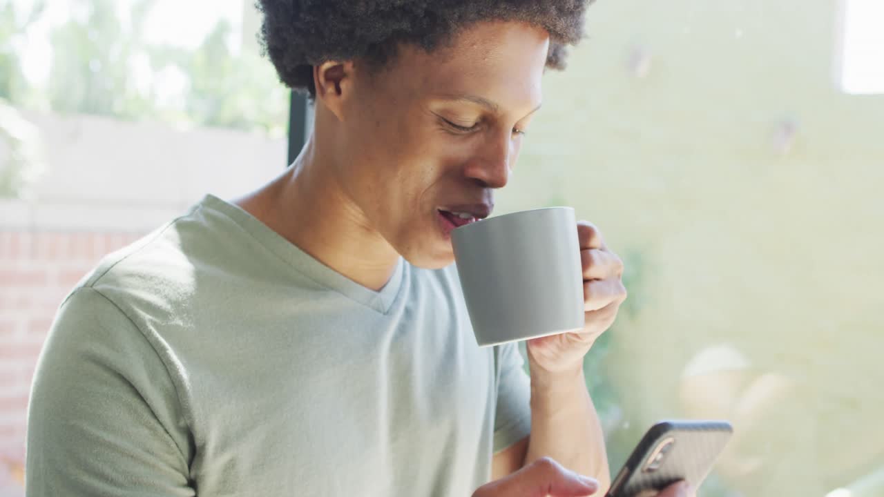 hombre afroamericano usando un teléfono inteligente y bebiendo café en casa