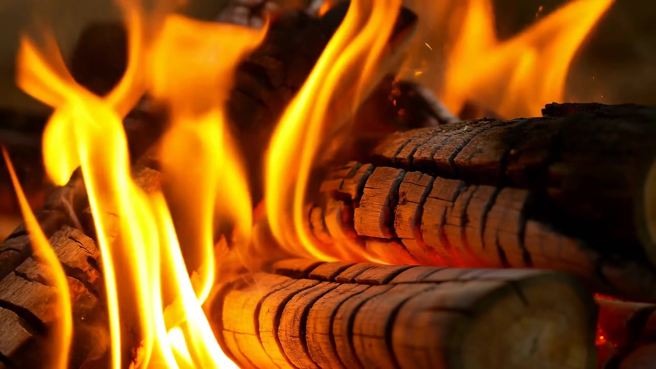 Lavt nærbillede af stablet brænde, der brænder aktivt, mens høje gul-orange flammer stiger og krøller over forkullede træstammer, glødende gløder, stigende varme, røg, rig tekstur og rustik varme