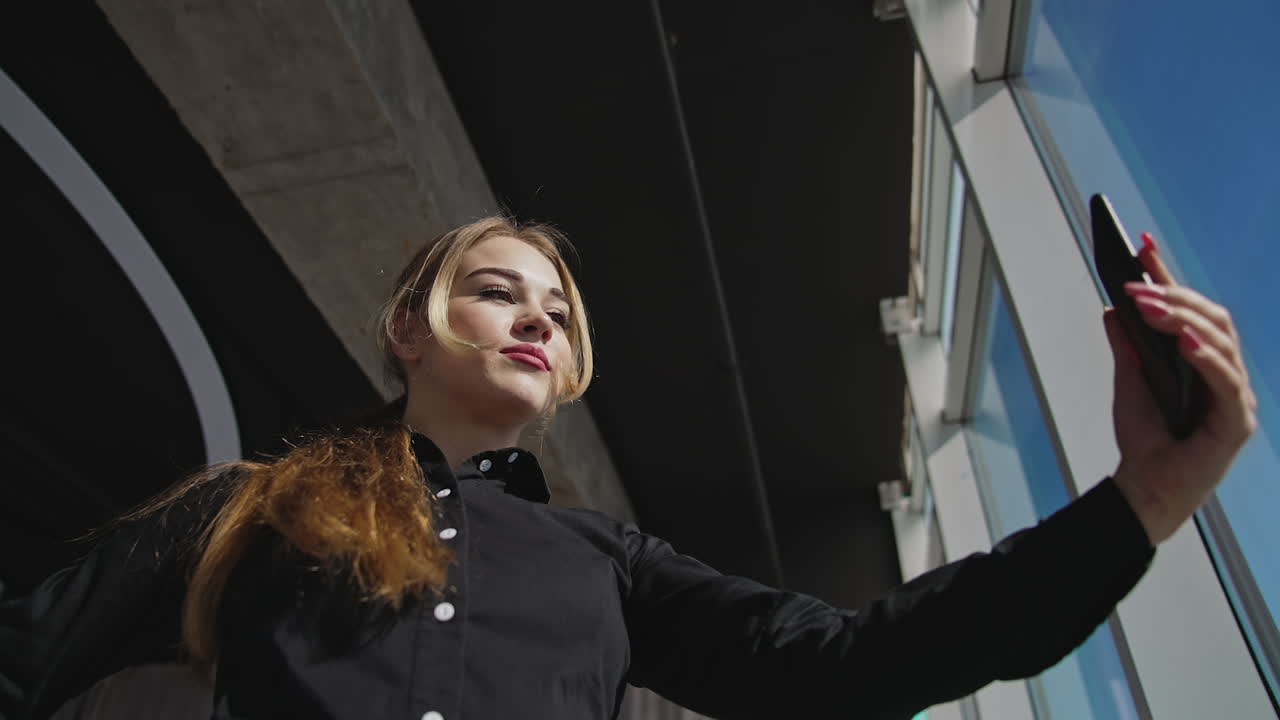 Beautiful lady taking video on her smartphone. Taking selfie near the sunny panoramic window. Low angle view.
