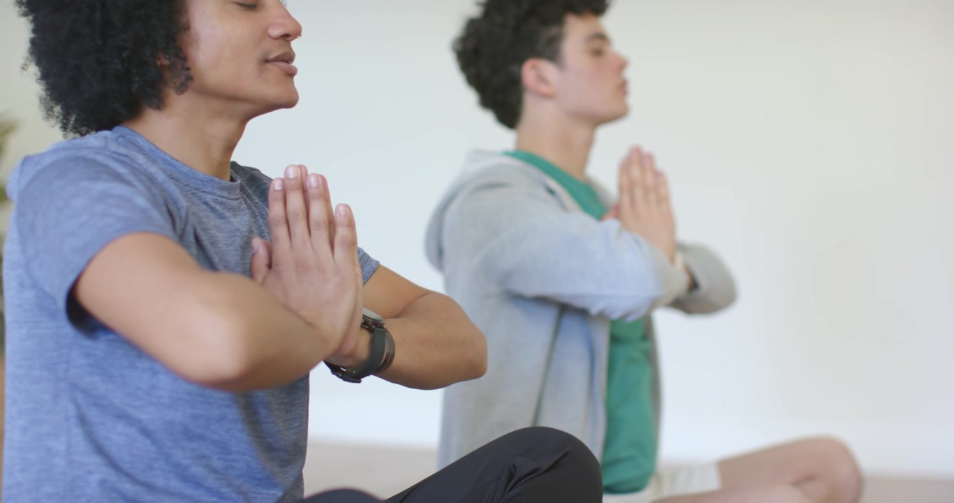 Two men sitting on studio mats following cue, shifting hands on knees, breathing, one wearing watch