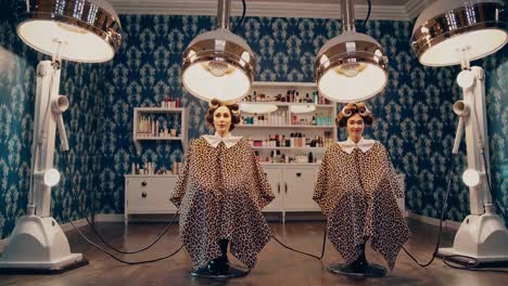 twin sisters with curlers in their hair wearing leopard print capes sit in a vintage hair salon, waiting for their hair treatment under bright lamps