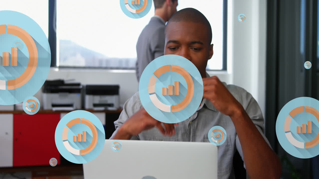 Animation of multiple graph icons over tired african american man looking at laptop in office