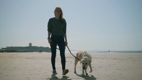 Lächelnde-Schöne-Frau-Mit-Brille,-Die-Mit-Hund-Am-Strand-Spaziert.