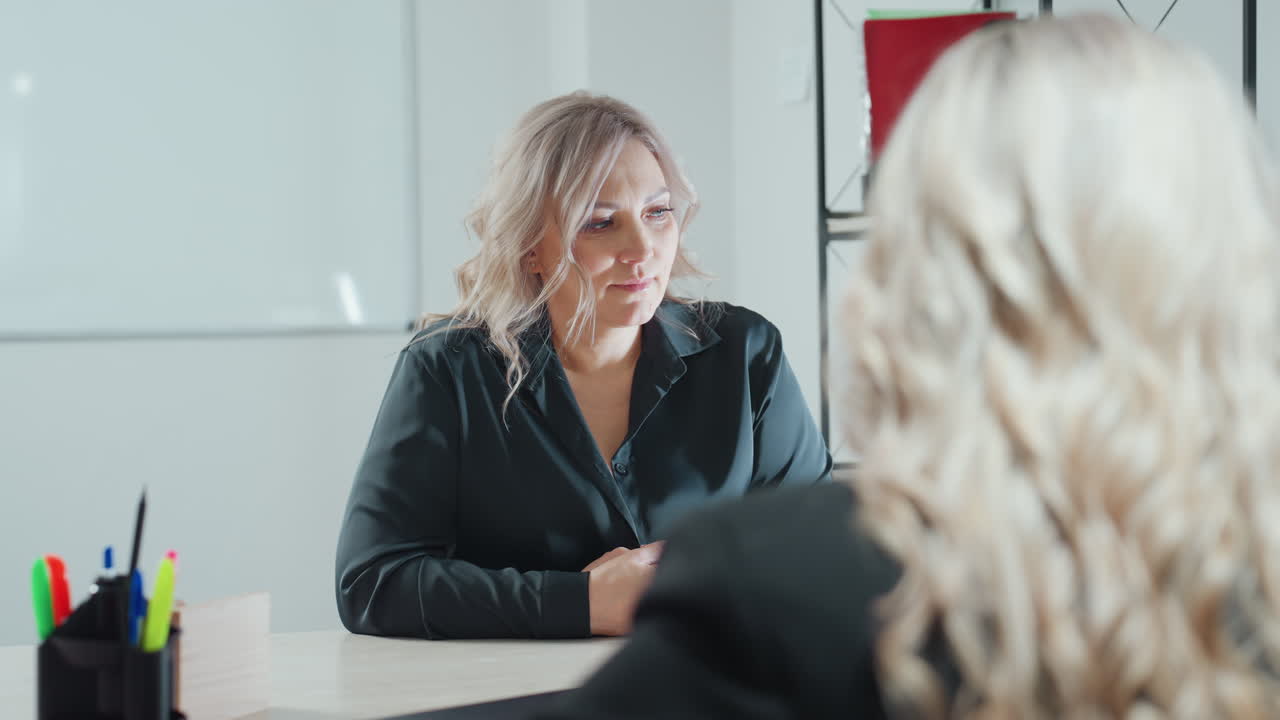 client paying attention to what ceo is telling her seated across desk with hands clasped in brightly lit office featuring indoor plants shelf and stationery holder showing engaged expression