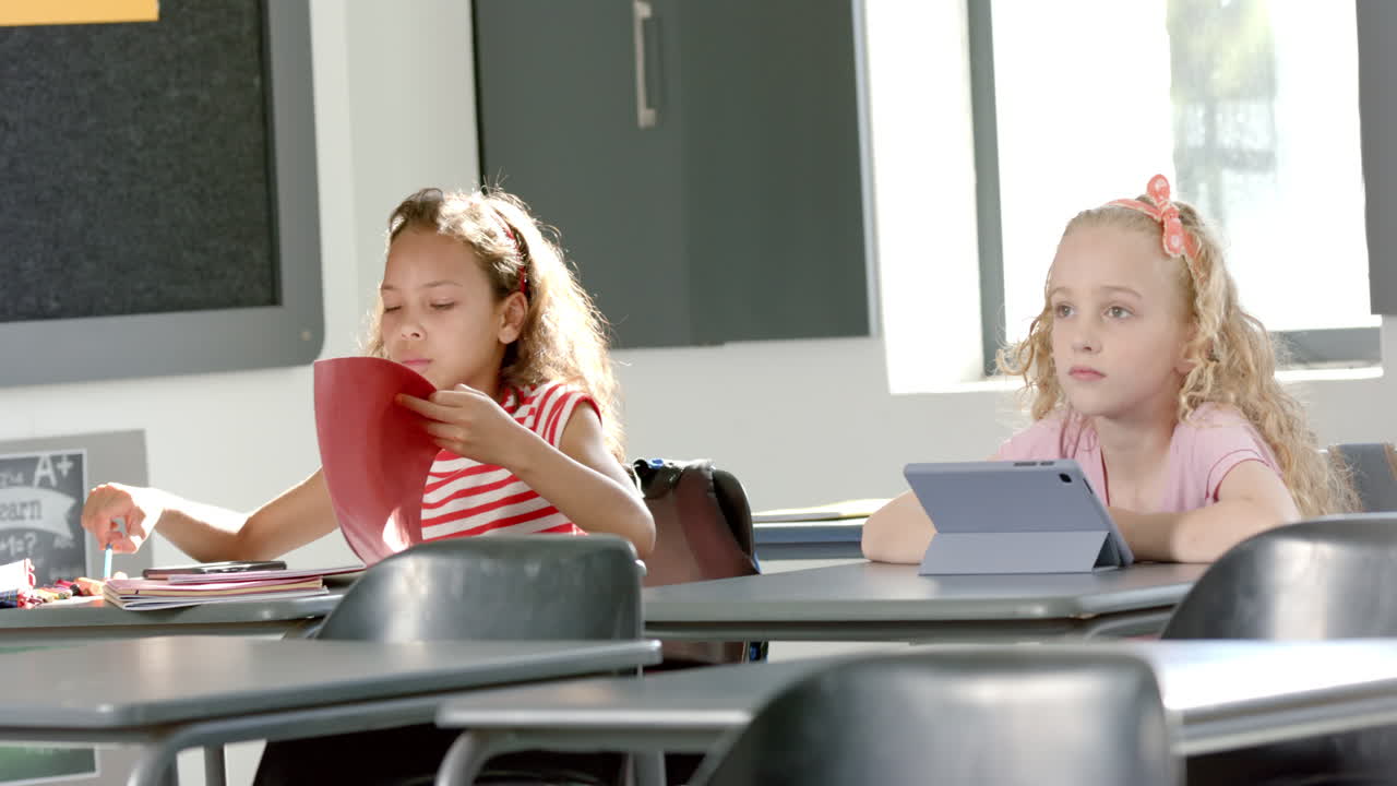 Biracial girl focused on her studies in a school classroom while Caucasian girl looks on curiously