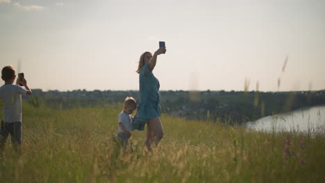 a woman in a blue dress holds her younger son and a phone, capturing moments in a grassy field during the golden hour. her older son is also holding a phone