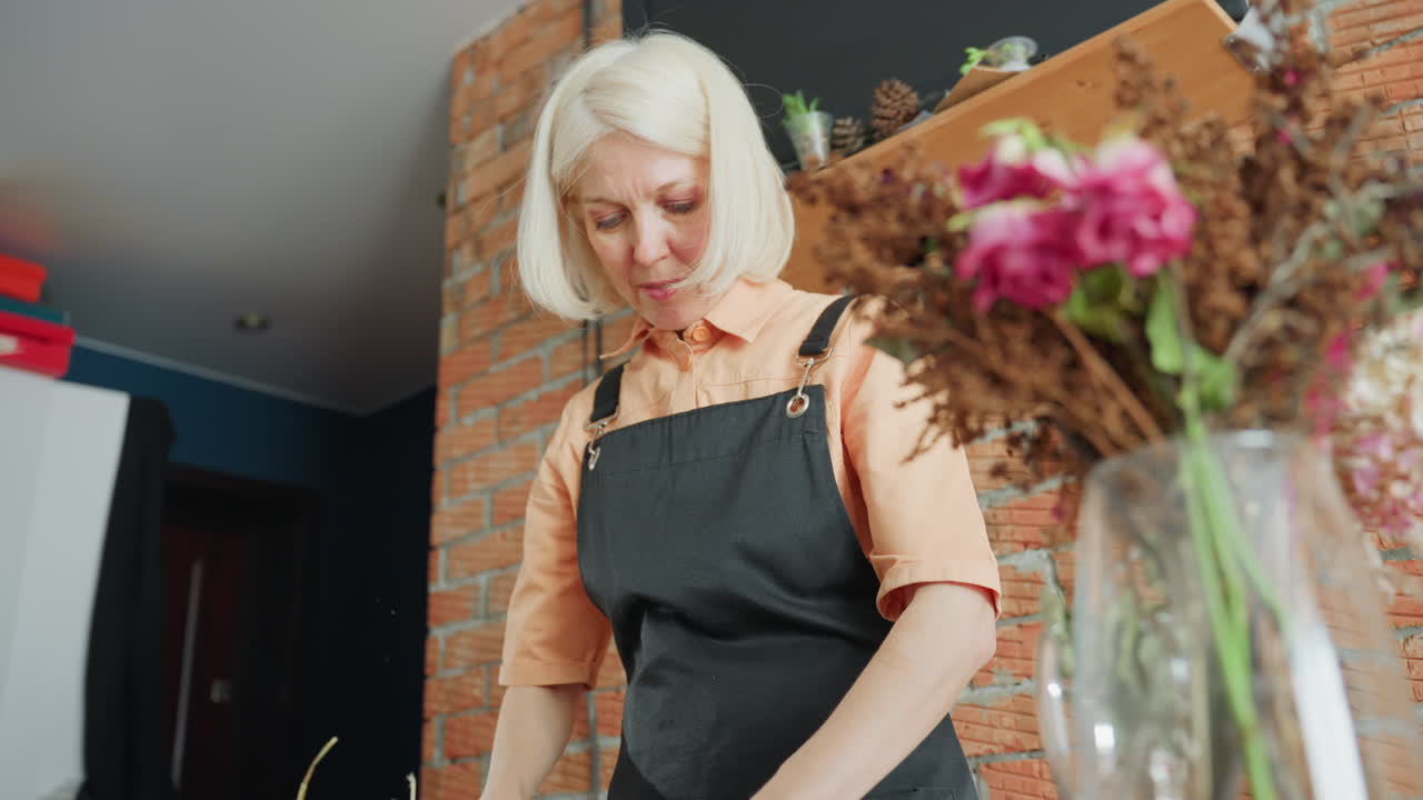 Senior woman florist in black apron working in workshop with blurred face in background while dried and fresh flowers including pink roses and brown stems stand in clear glass vase in foreground