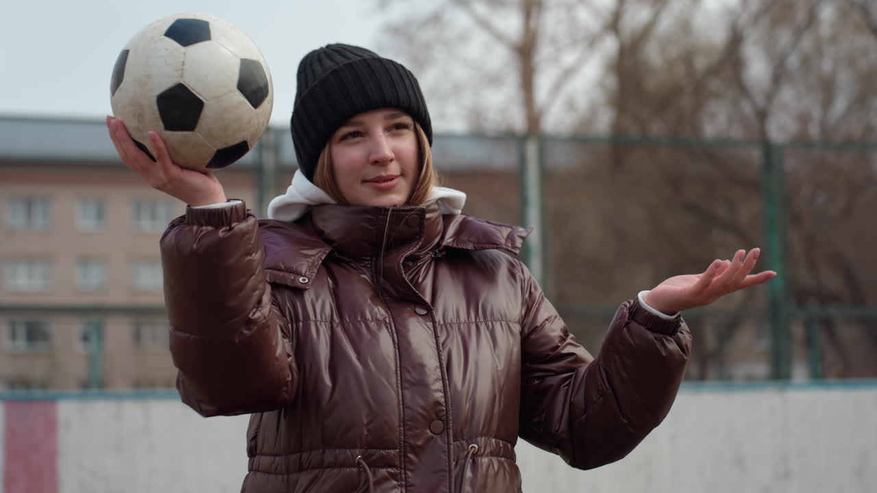 Hvit jente sjonglerer fotball på utendørsbane, lue og boblejakke, smiler mens hun øver på kast, venn i grønn jakke ser på, lavt vinterlys, ærlig atletisk lek og glede