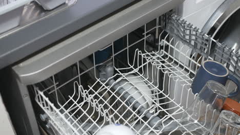 loading a dishwasher rack with mugs and glassware. the open dishwasher is filled with assorted clean kitchen items
