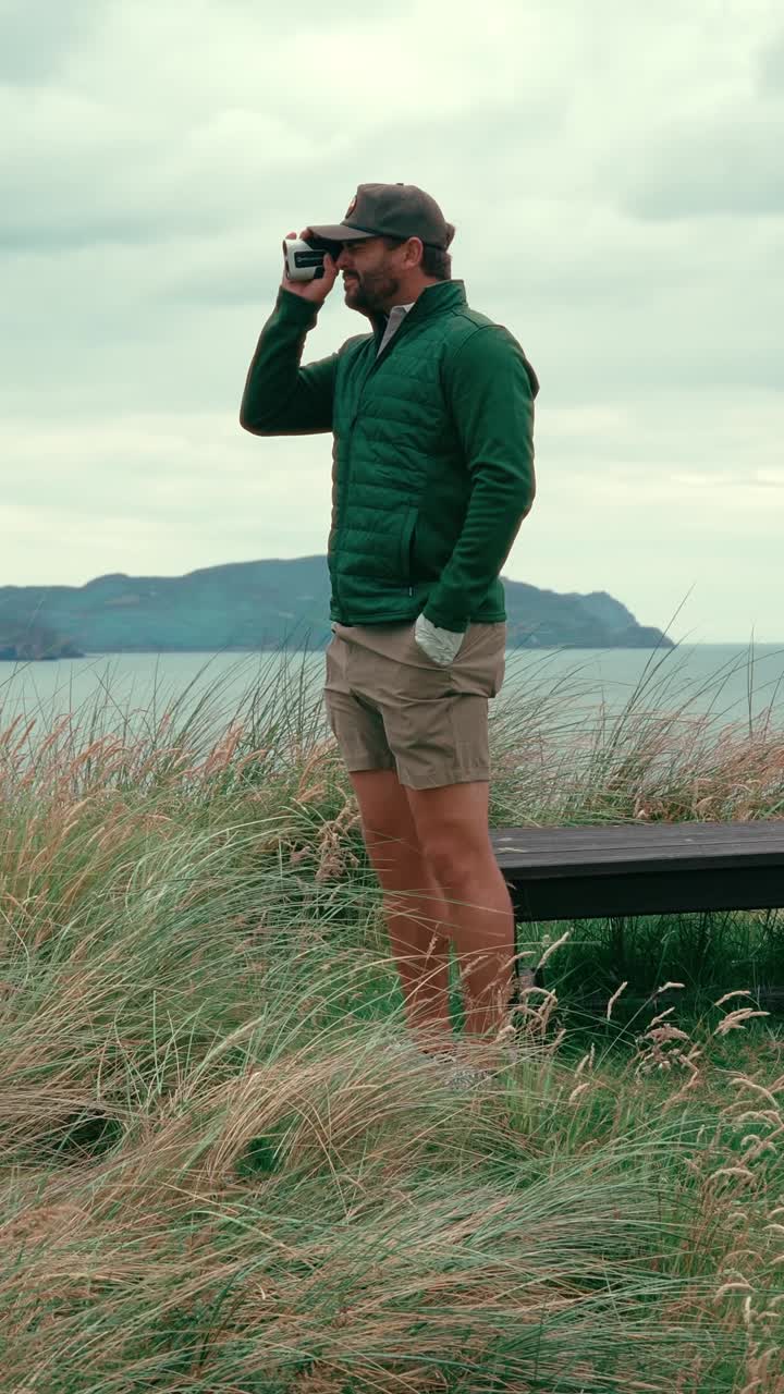 Vertical shot of male golfer standing in rough using range finder to get accurate yardage to pin on coastal Ireland links golf course on windy day