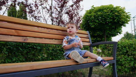 beautiful caucasian baby sitting on the bench in summer. kid holds a bottle of water in hands. low angle view.