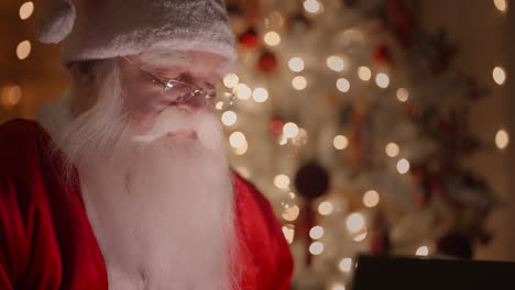 vue latérale en gros plan d'un vrai père noël travaillant avec un ordinateur portable la nuit avec des lunettes à la lumière des lumières de noël sur le fond d'un arbre de noël. travail à distance