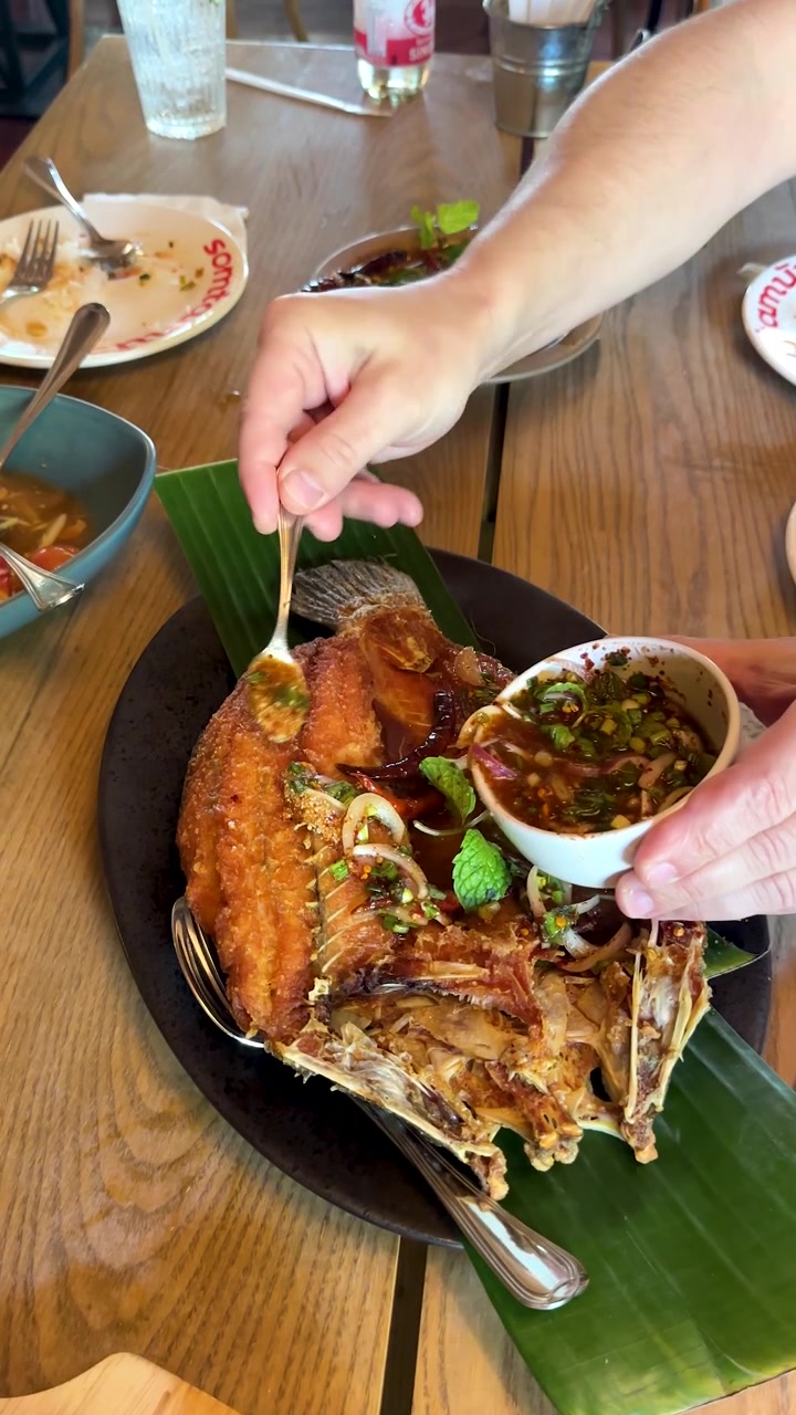 Tangan menuangkan saus cabai herbal pedas ke atas ikan goreng renyah di restoran Thailand, dengan cahaya alami.