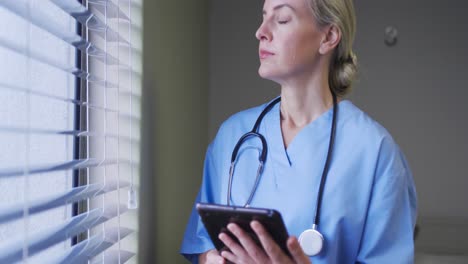 Portrait-of-caucasian-female-doctor-holding-tablet-looking-through-the-window-in-hospital-room