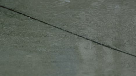 Gotas-De-Primer-Plano-En-Cámara-Lenta-De-Lluvia-De-Verano-O-Otoño-De-Agua-Caen-Sobre-El-Asfalto-En-La-Ciudad-Convirtiéndose-En-Un-Gran-Charco,-Inundando-La-Calle