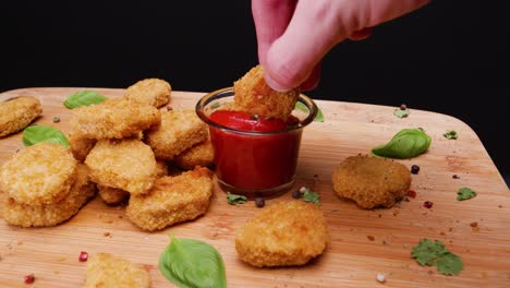 Dipping-vegetarian-vegan-chicken-nuggets-in-sauce-on-a-bamboo-cutting-board-with-basil-leaves-and-peppercorns-for-garnish,-black-background,-static-shot