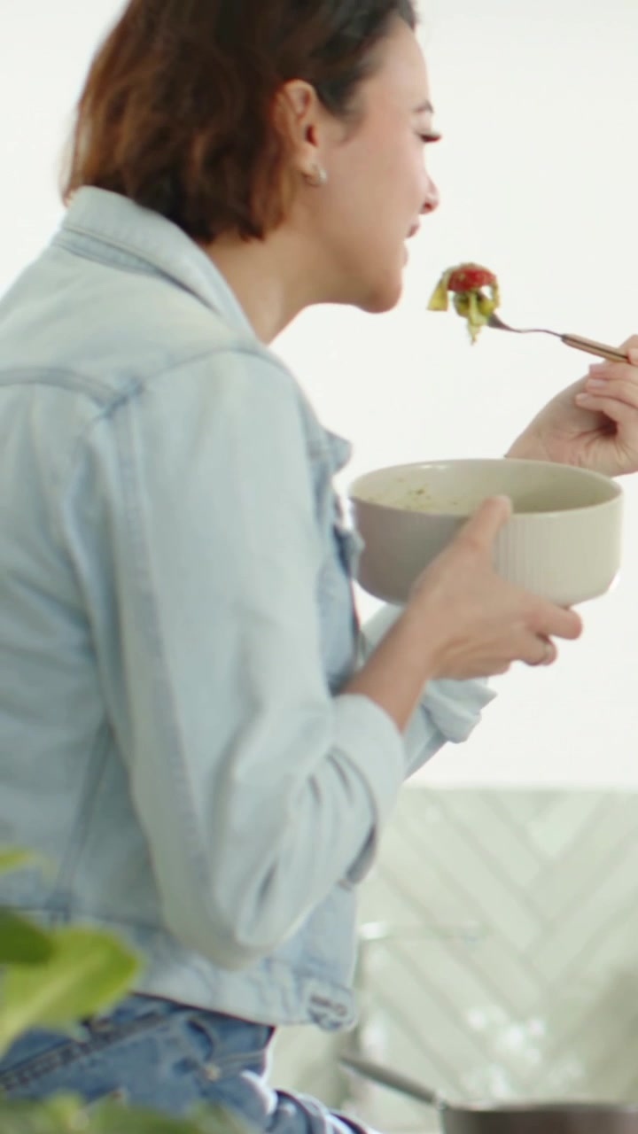 세로 영상: 집 카운터에서 포크를 들고 세라믹 그릇에 담긴 음식을 먹는 여성
