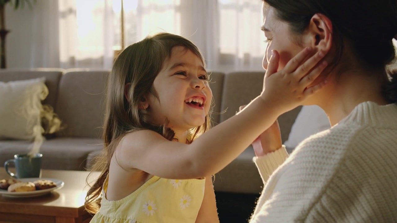 黄色いドレスを着た笑顔の子供が、ソファに座る母親に手を伸ばし、マグカップのそばで優しく頬に触れている様子