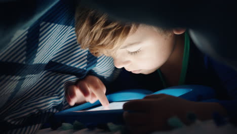 happy little boy using digital tablet computer under blanket enjoying learning on touchscreen technology playing games having fun at bedtime