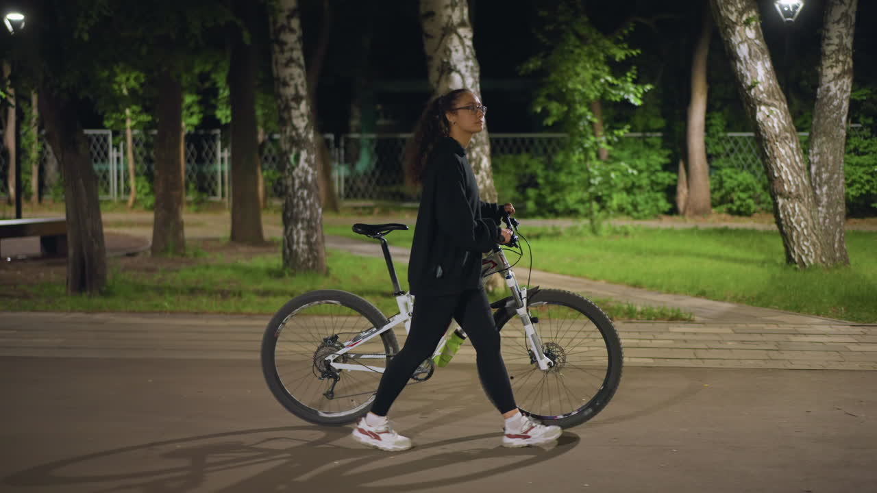 Women Cycling Confidently, Night Walk Under Shaded Branches, Female Explorer Journeys Through Dusky Leafy Avenues, Woman Confidently Rides Her Bicycle As Night Falls Under Lush Green Trees