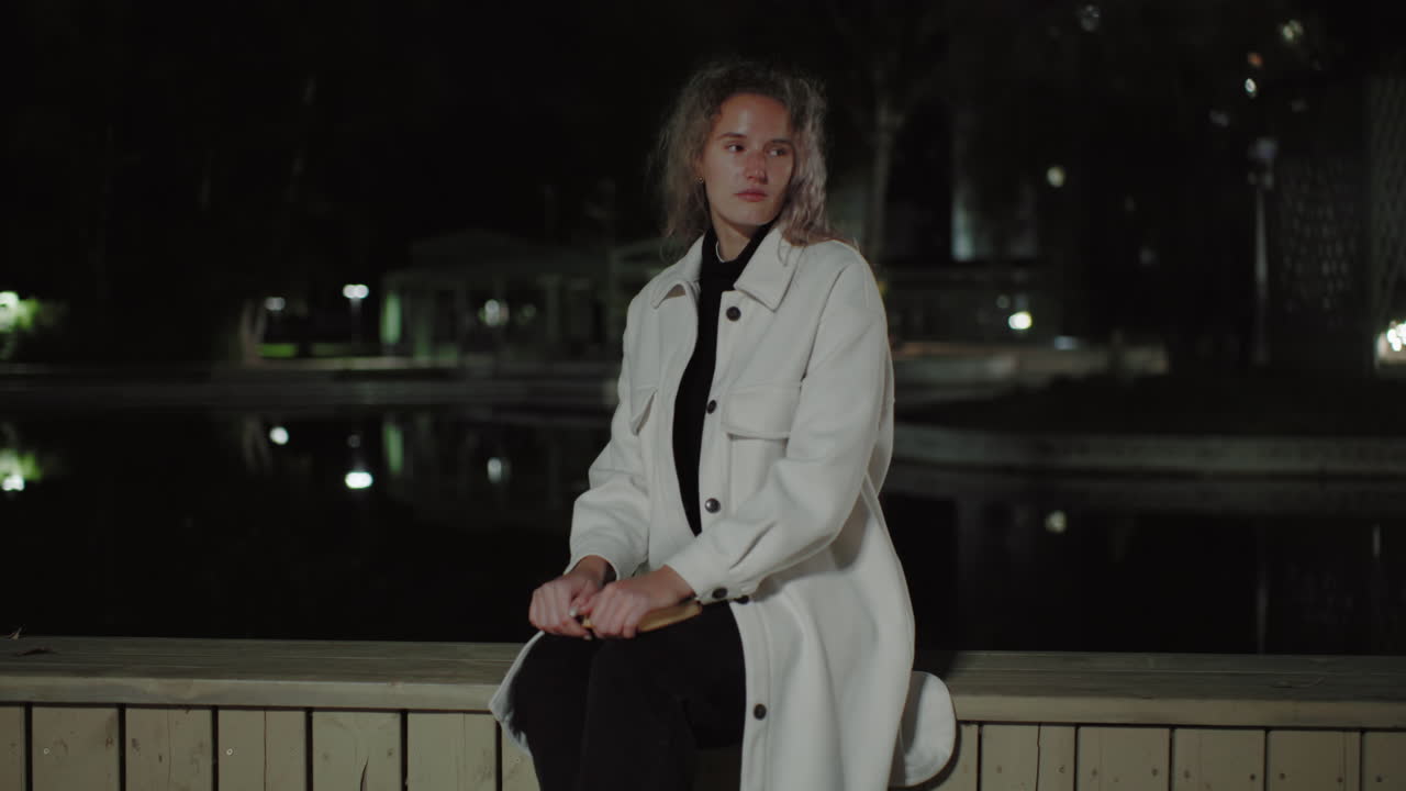 Woman in white coat sits on wooden fence by calm water at night, city lights reflecting gently, capturing peaceful scene with subtle architecture in background