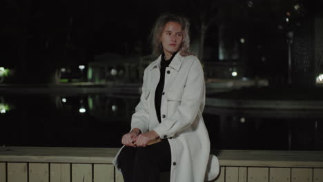 woman in white coat sits on wooden fence by calm water at night, city lights reflecting gently, capturing peaceful scene with subtle architecture in background