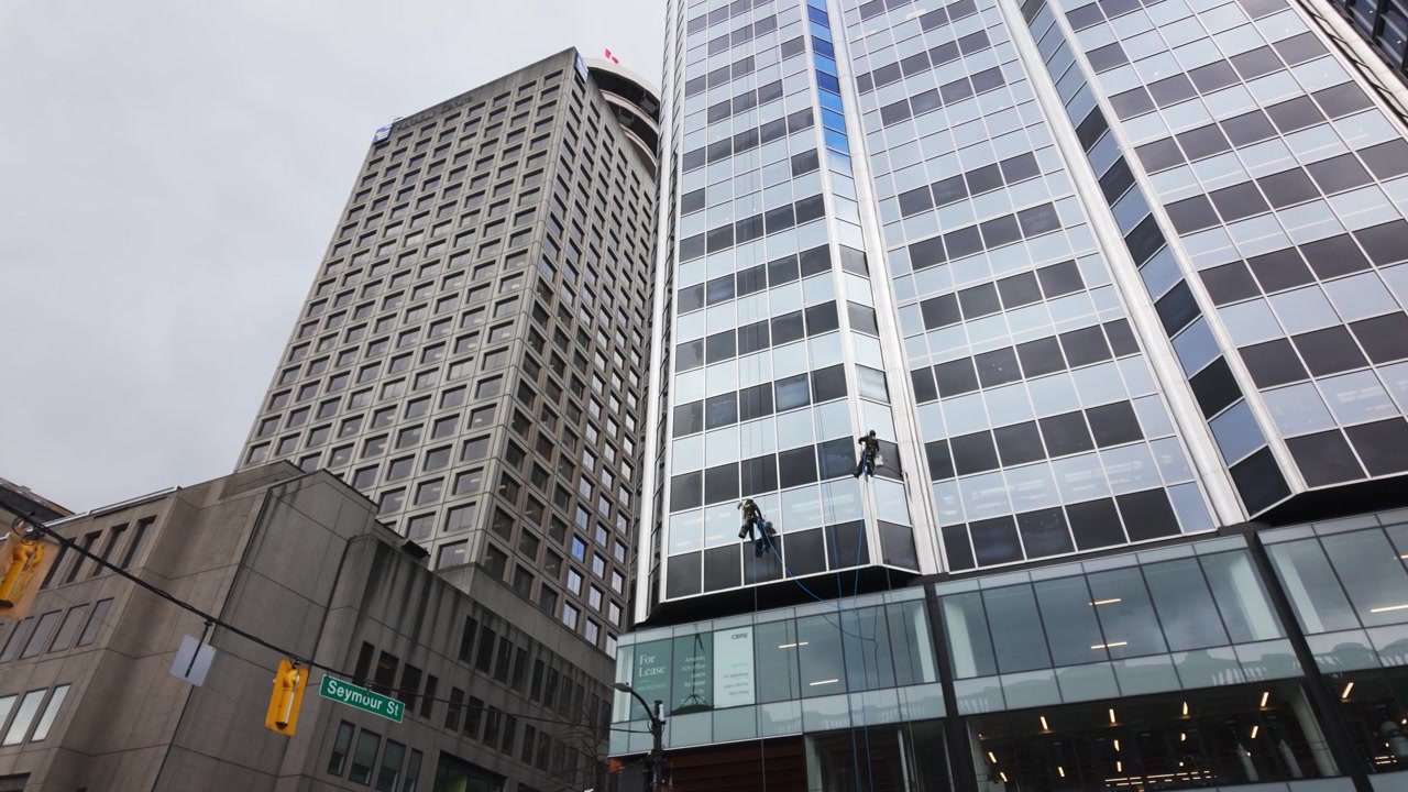 Dos limpiadores de ventanas están suspendidos por cuerdas mientras limpian un rascacielos de cristal en el centro de Vancouver. Con un telón de fondo de edificios de oficinas modernos e históricos en Seymour Street