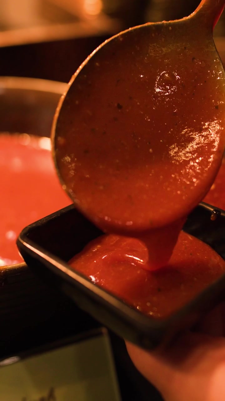 Una mano versa con un mestolo salsa rossa calda in una ciotola nera a un buffet, primo piano, illuminazione calda e soffusa