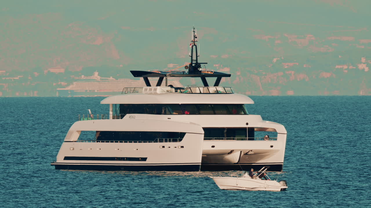 Un moderno yacht catamarano bianco ancorato su acque cristalline con una città costiera sullo sfondo