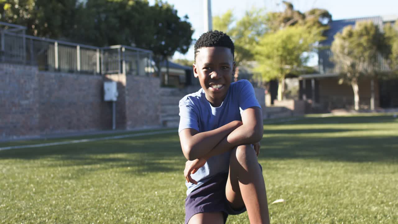Joven africano arrodillado sobre el césped con una camiseta azul claro, respondiendo a la cámara y sonriendo