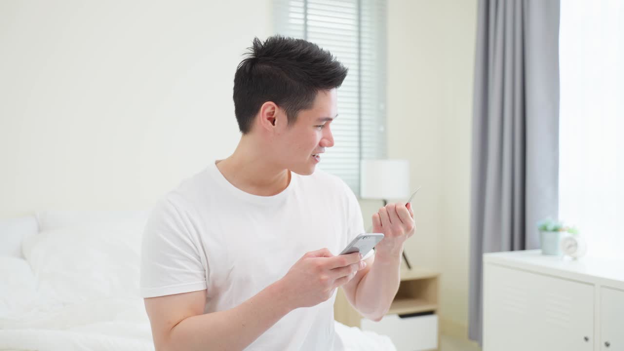 아시안 매력적인 남성은 아침에 집에서 침대에서 온라인 쇼핑을 즐긴다. 예쁜 남자는 침실에 누워있고, 신용카드를 들고 은행에서 온라인 지불을 할 수 있습니다.