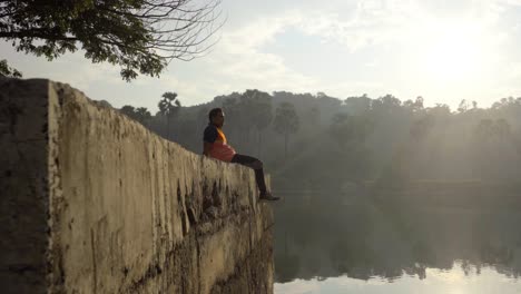 Junge-Sitzgelegenheiten-Auf-Einem-Dock-Am-See-Entspannt-Genießen-Die-Aussicht-In-Sonnenaufgang-Morgen-Junge-Mann-Wasser-See-Revil-Erschossen-Reflexion-Im-Wasser