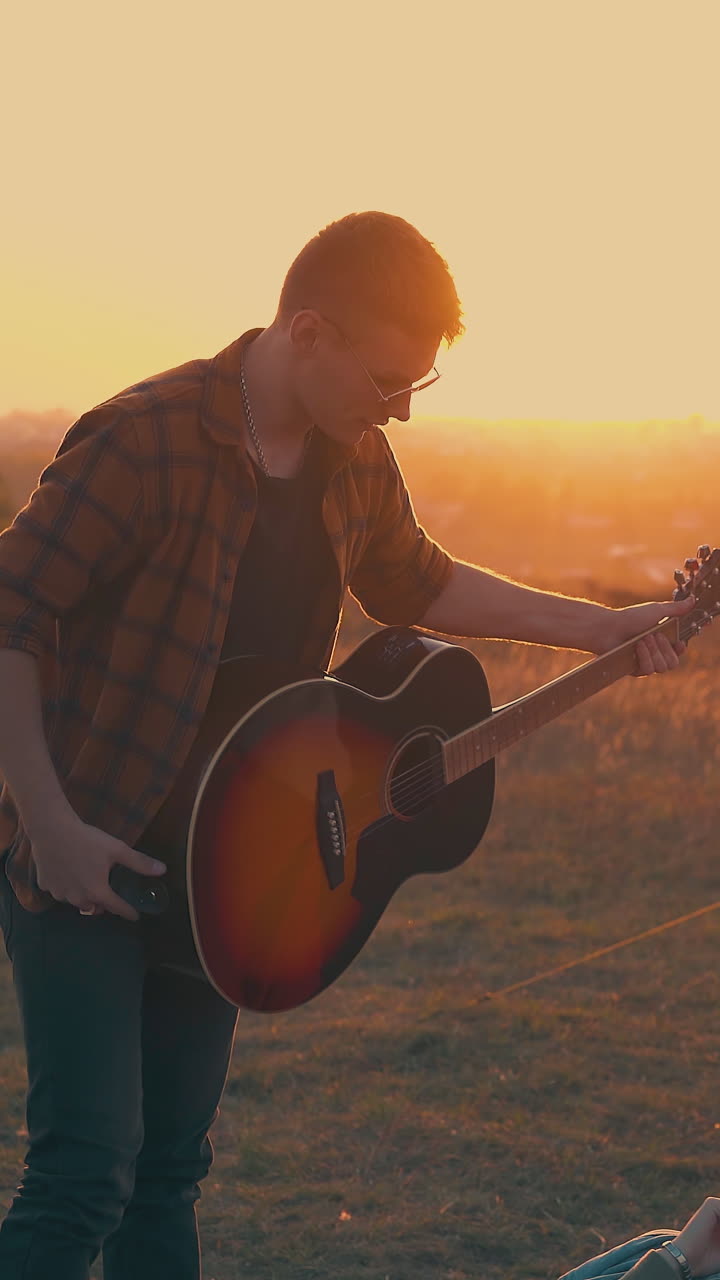 happy guy in red checkered shirt plays guitar to friends sitting near blue tent in warm autumn evening at back sunlight