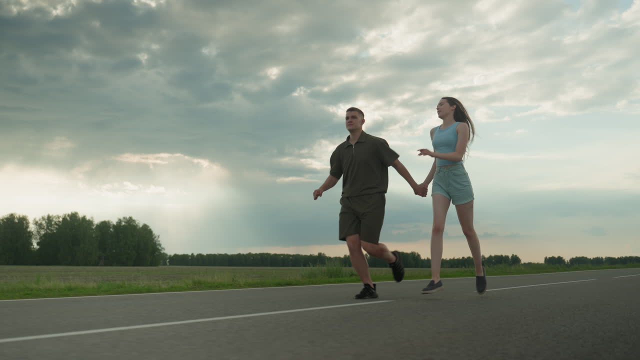 rear view of couple holding hands as man in olive shirt runs ahead while woman in blue tank and denim shorts chases him with hair flowing on rural road under dramatic cloudy sky at golden hour