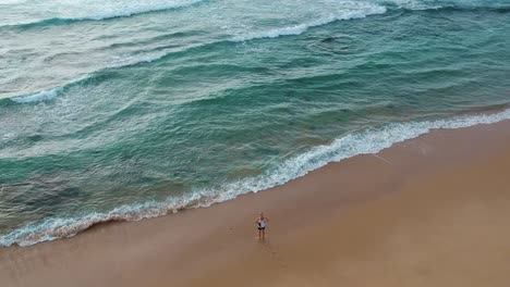 Chica-Sola-En-Una-Playa-Mirando-La-Hermosa-Vista-Aérea-En-ángulo-Del-Agua-Azul-Del-Océano