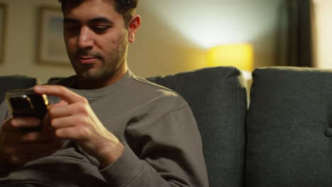 joven preocupado pasando la noche en casa sentado en el sofá con el teléfono móvil desplazándose a través de internet o medios sociales de transmisión de contenido 2