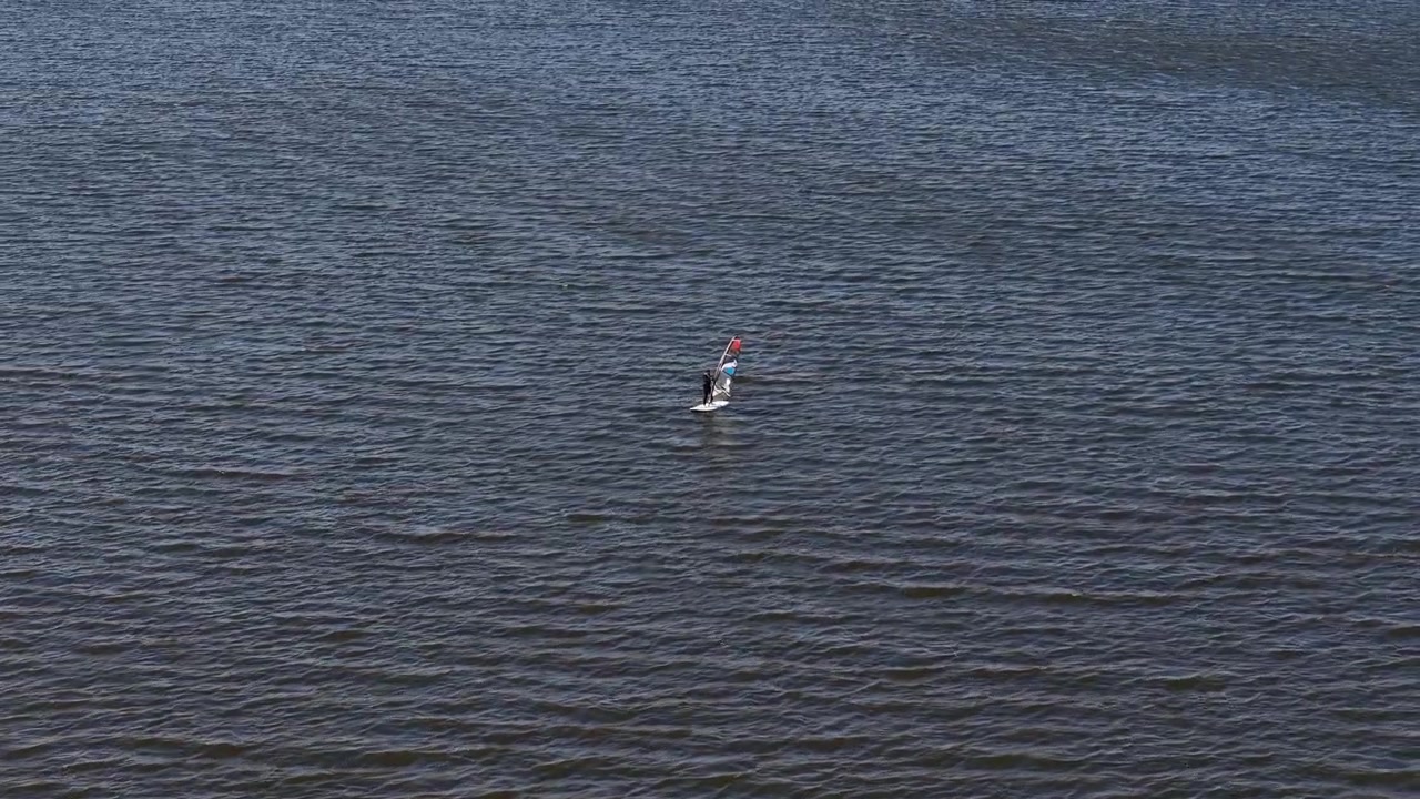 Windsurfer sails across dark choppy water on a colorful board and sail, with wide open water creating generous copy space around the subject.