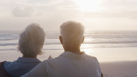 Vista-Trasera-De-Una-Pareja-Mayor-En-La-Playa-Viendo-La-Puesta-De-Sol-Sobre-El-Océano