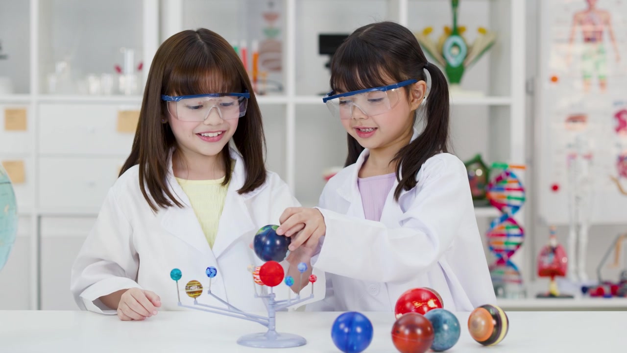 Two schoolgirls wearing safety goggles examine a planetary model in a bright science classroom.