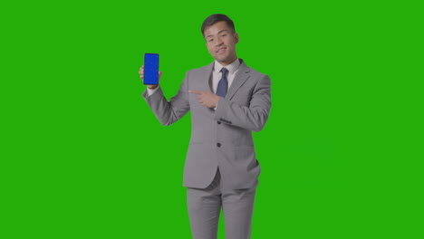 studio shot of smiling businessman in suit holding blue screen mobile phone against green screen 3