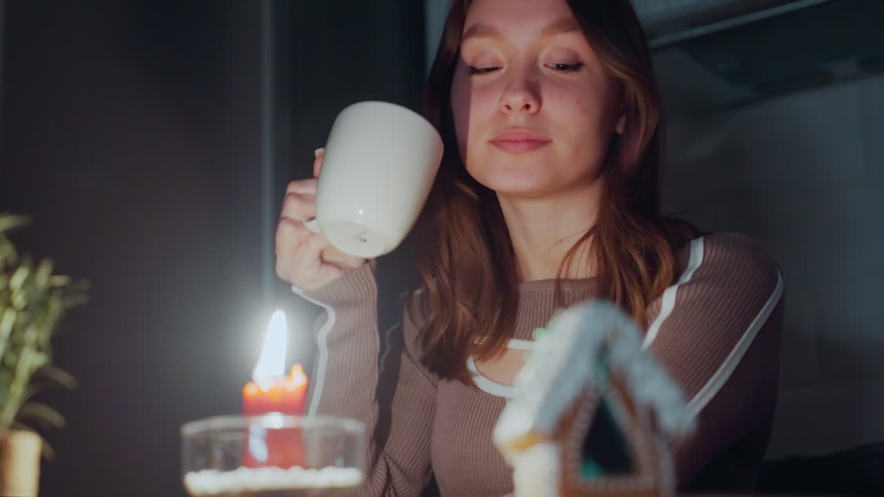 primer plano de una vela roja ardiendo al lado de una casa de pan de jengibre con glaseado blanco, una mujer bebe té, bajando la taza mientras descansa la cabeza sobre su mandíbula, mirando pensativo a la casa de galletas
