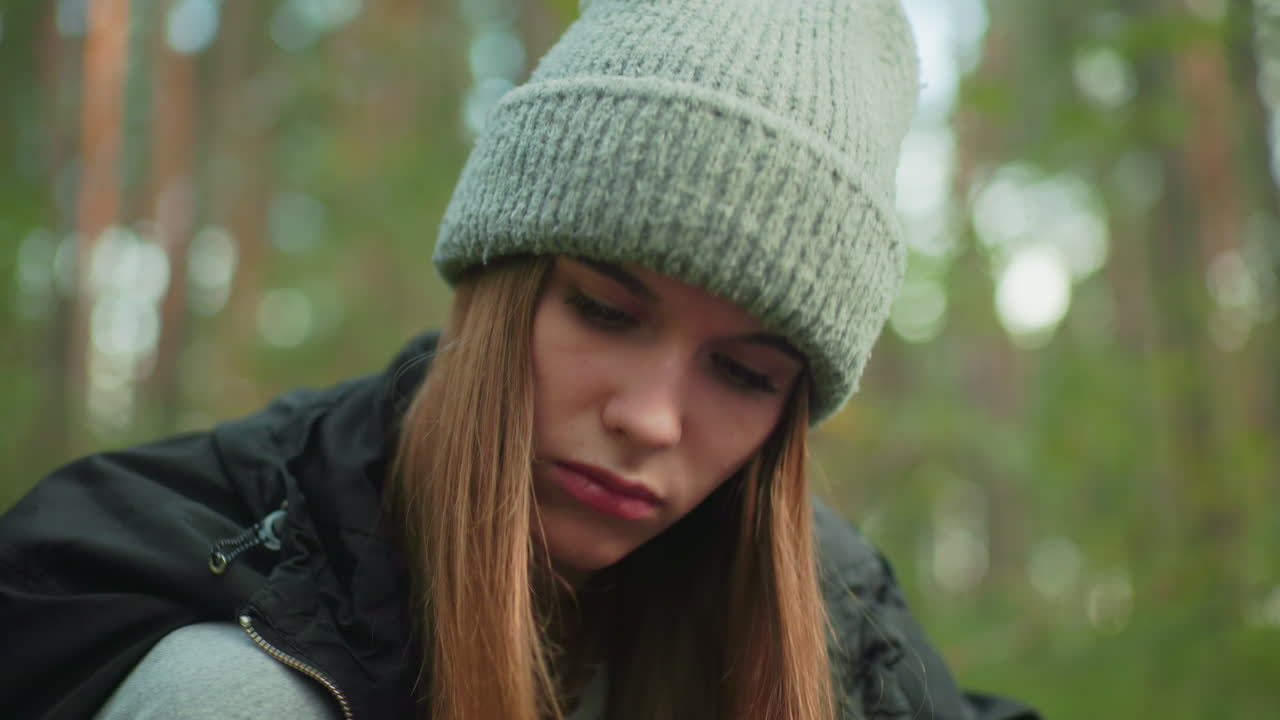 close up of lady in gray beanie and black jacket concentrating intensely as she hits something with wood in background surrounded by green forest, face focused with determination and intent