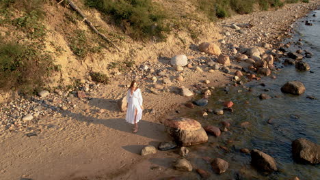 Carefree-Woman-in-White-Long-Summer-Dress-Walking-Barefoot-on-Wild-Rocky-Sea-Shore-By-the-Water-at-Sunrise---Aerial-High-Angle-Push-Back